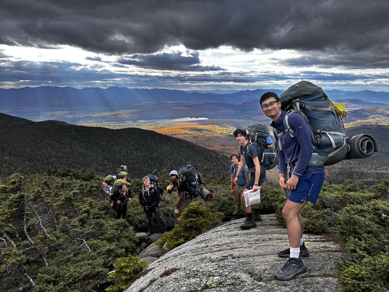 A group of hikers is making their way up a rocky mountain trail. The hikers are wearing backpacks and hiking gear. The sky is cloudy and the landscape is green and mountainous. The man in the foreground is smiling and looking at the camera. He has a very large backpack. 